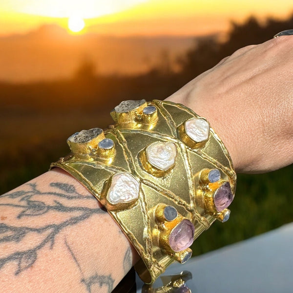 A person's wrist wearing a gold-plated brass cuff bracelet featuring mother-of-pearl crystals and other stones, with a sunset in the background.