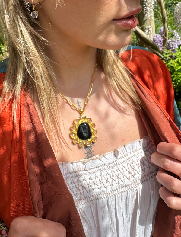 A person wearing an orange jacket and a white top with a gold-plated silver pendant necklace featuring a black agate druzy at the center, surrounded by citrine crystals in a blooming formation.