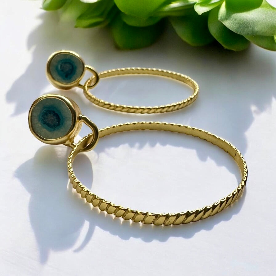 Gold hoop earrings with green gemstones on a white surface with green leaves in the background
