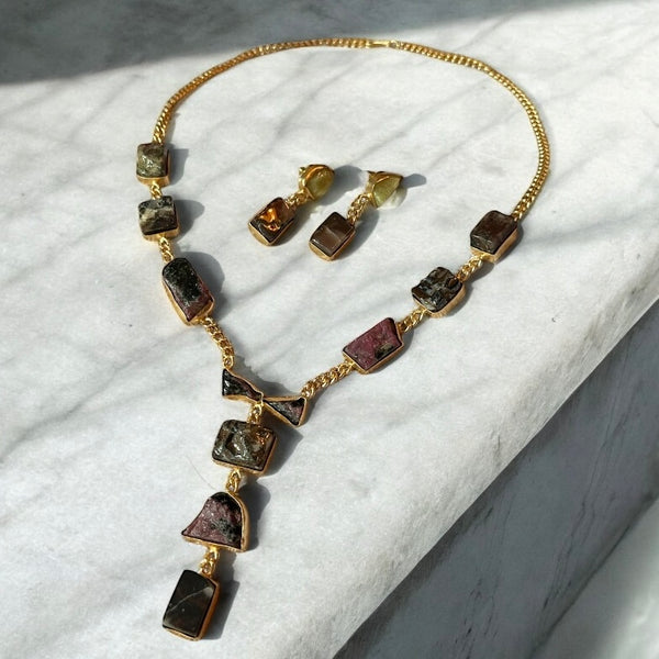 A gold-plated brass necklace set with raw cut smoky quartz and fuschite charms, displayed on a marble surface.